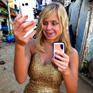 blond self-indulgent instagram valley girl in a prom dress taking a selfie with a gold iphone in a favela surrounded by poverty and filth
